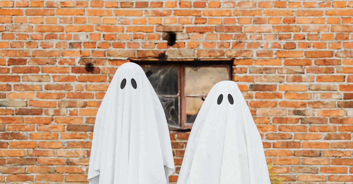 Two ghostly figures in white sheets stand against a rustic brick wall for Halloween.